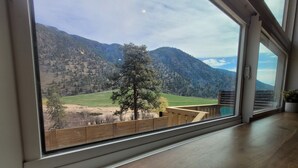 A room with a large window offering a view of a mountainous landscape, a wooden deck outside, and a potted plant on the windowsill.