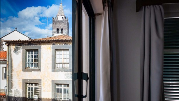 Une vue à travers la fenêtre d’un bâtiment doté d’un toit en tuiles, d’un clocher et d’une fenêtre ornée d’un rideau.