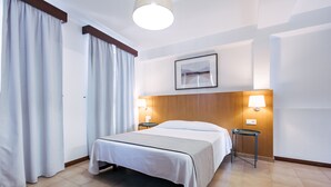 Une chambre d’hôtel avec un lit, deux tables de chevet, une lampe fixée au mur, un tableau encadré et une fenêtre avec des rideaux.