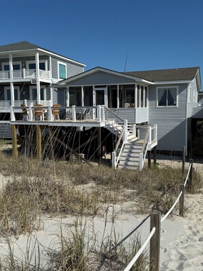 A beachfront house with a covered deck, a staircase leading to a second level, and a sandy beach in the foreground. - Amazing 3-bedroom beach house with an enchanting view, screen porch & deck (Oak Island)