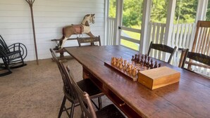 A wooden dining table with chess set, chairs, a rocking horse, and a pitchfork on a porch with white railings and a view of greenery.