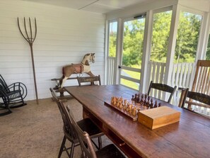 A wooden dining table with chess set, chairs, a rocking horse, and a pitchfork on a porch with white railings and a view of greenery.