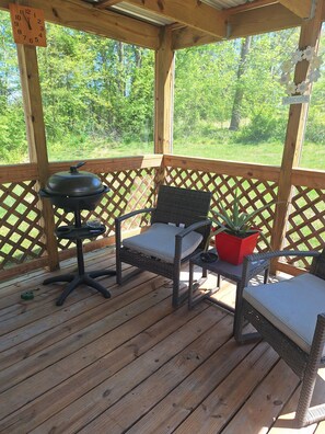 Une terrasse extérieure couverte, dotée d’une rambarde en treillis de bois, d’un barbecue et de deux fauteuils en osier avec des coussins.