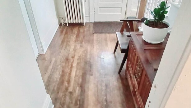 A hallway with wooden flooring, a white door, a radiator, a small table with a potted plant, and a staircase with white balusters.