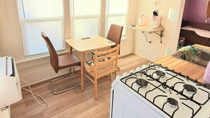 A compact kitchen with a gas stove, wooden dining table, and chairs. There are three windows with blinds, a small shelf with kitchen items, and a visible power outlet. - Windrose: Tranquil Pets-OK Soak Tub Suite Near ISU (Ames)