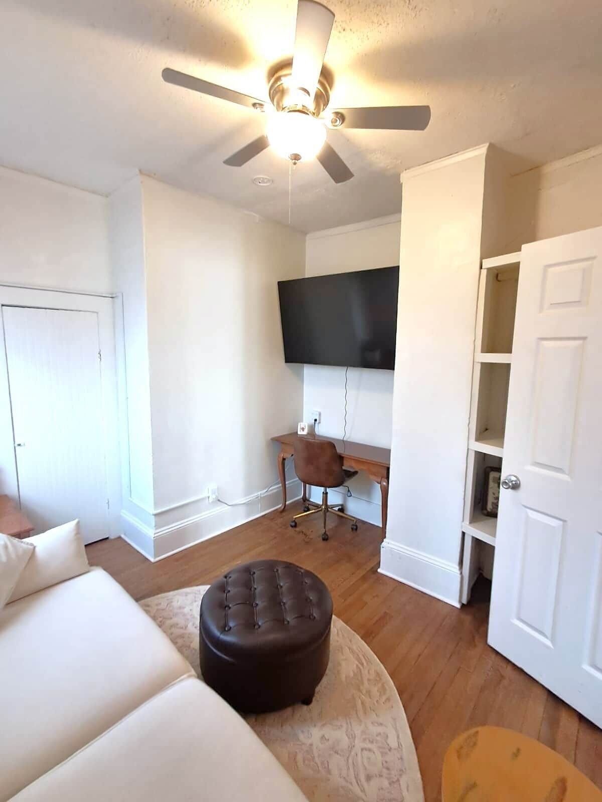 Una habitación con ventilador de techo, televisión de pantalla plana montada en la pared, un escritorio de madera, un reposapiés de cuero marrón y un sofá blanco.