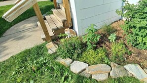 A wooden staircase leading to a white building, surrounded by a garden with various plants and rocks. - Windrose: Tranquil Pets-OK Soak Tub Suite Near ISU (Ames)