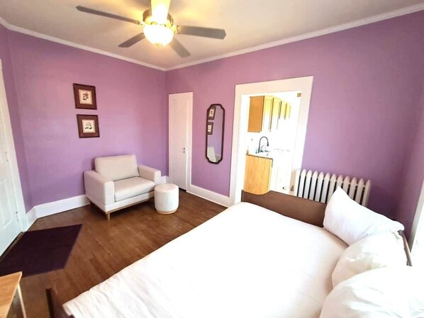 Une chambre à coucher avec un lit, un canapé blanc, une petite table ronde, un ventilateur de plafond et une fenêtre donnant sur un immeuble.
