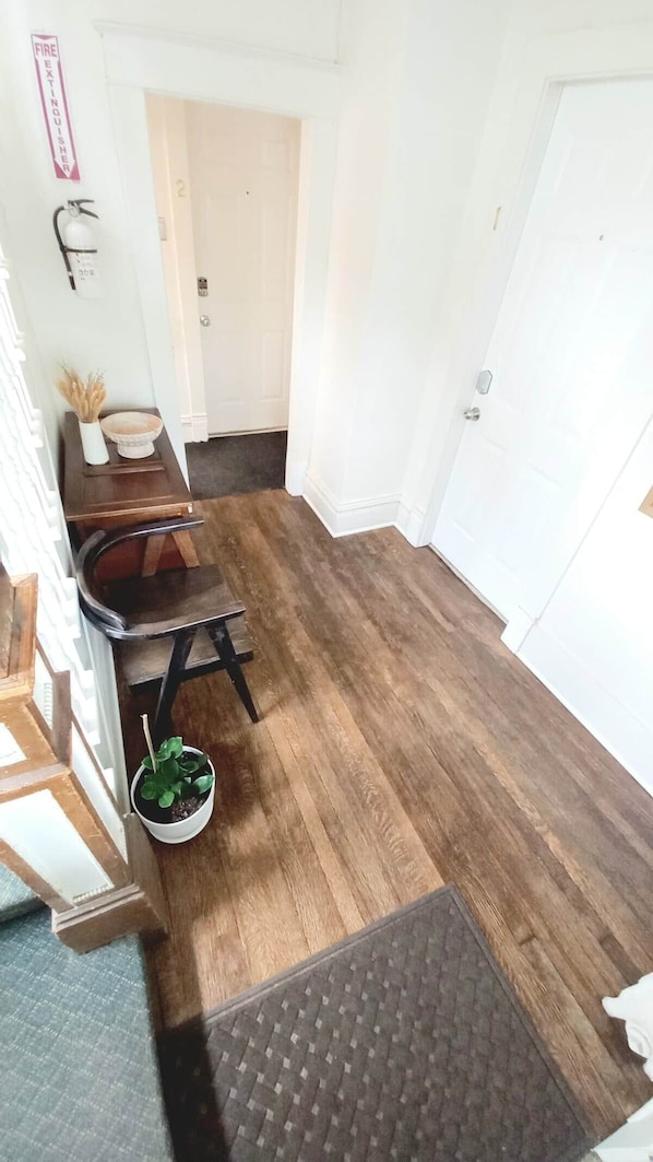 A hallway with a wooden floor, a small table with a plant, a chair, and a door with a number.