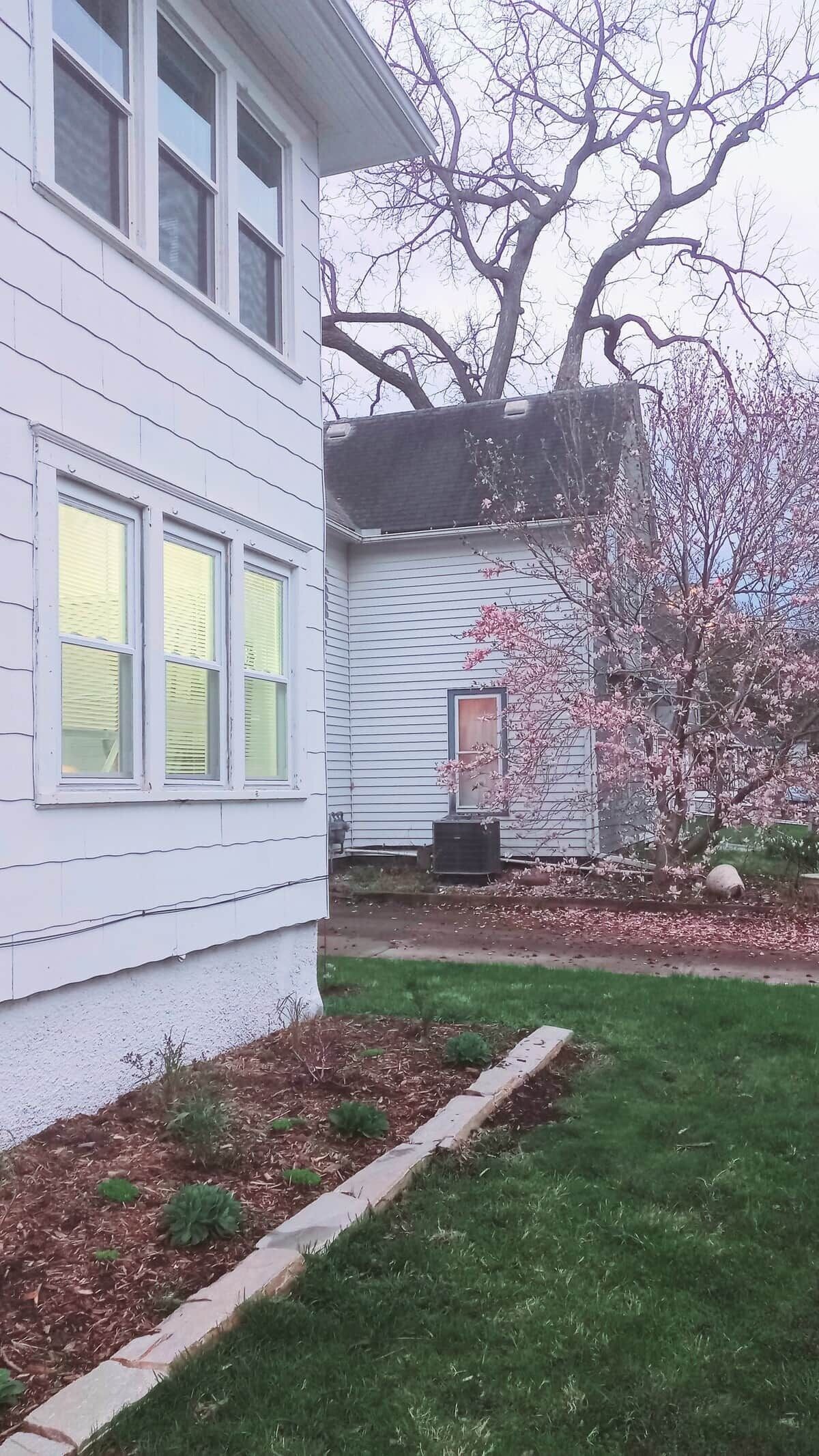Una casa de dos pisos con un jardín delantero, un pequeño árbol con flores rosadas y un equipo de aire acondicionado visible.