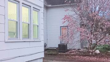 Una casa de dos pisos con un jardín delantero, un pequeño árbol con flores rosadas y un equipo de aire acondicionado visible.