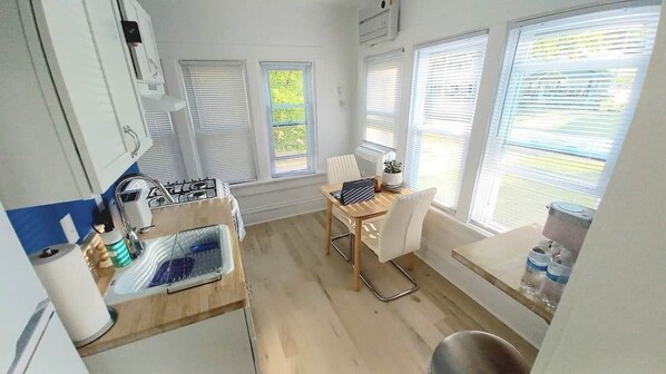 A compact kitchen with a sink, a small table, and a chair. There are windows with blinds, a water dispenser, and a trash bin.