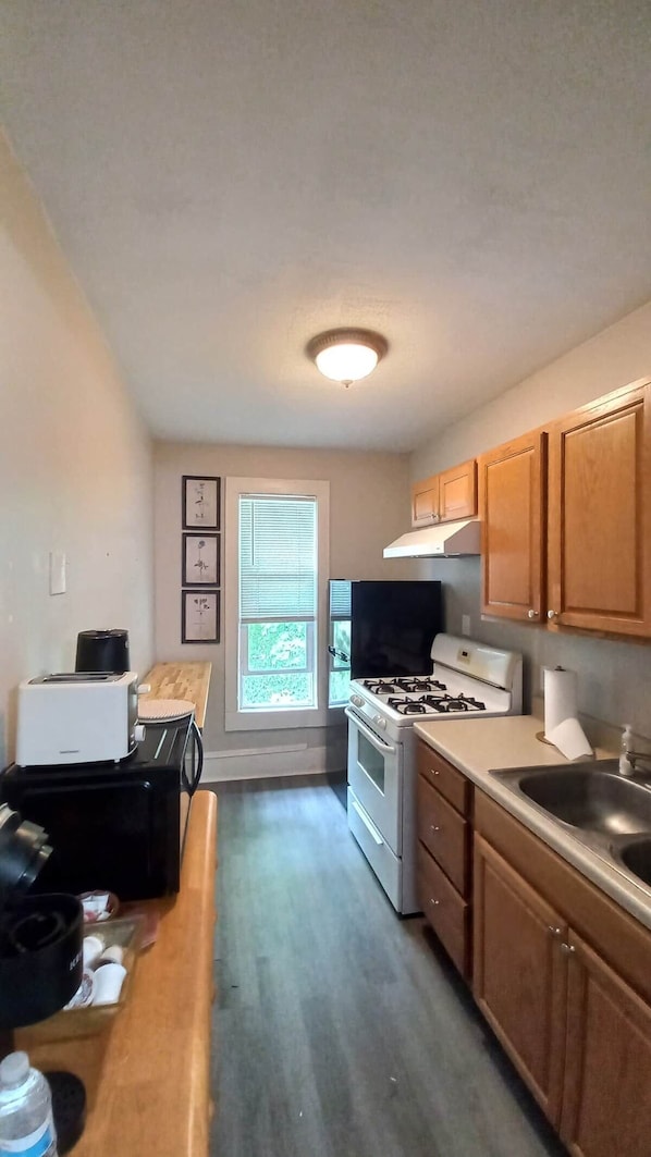 A kitchen with a gas stove, oven, sink, and wooden cabinets. There is a window with blinds, a toaster, and a refrigerator.