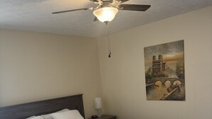 A bedroom with a bed, a ceiling fan, a nightstand, a wall clock, and a painting.
