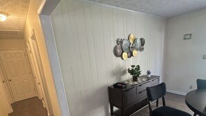 A dining area with a wooden floor, a dining table, two chairs, a small side table with a plant, and a wall-mounted decorative plate arrangement.
