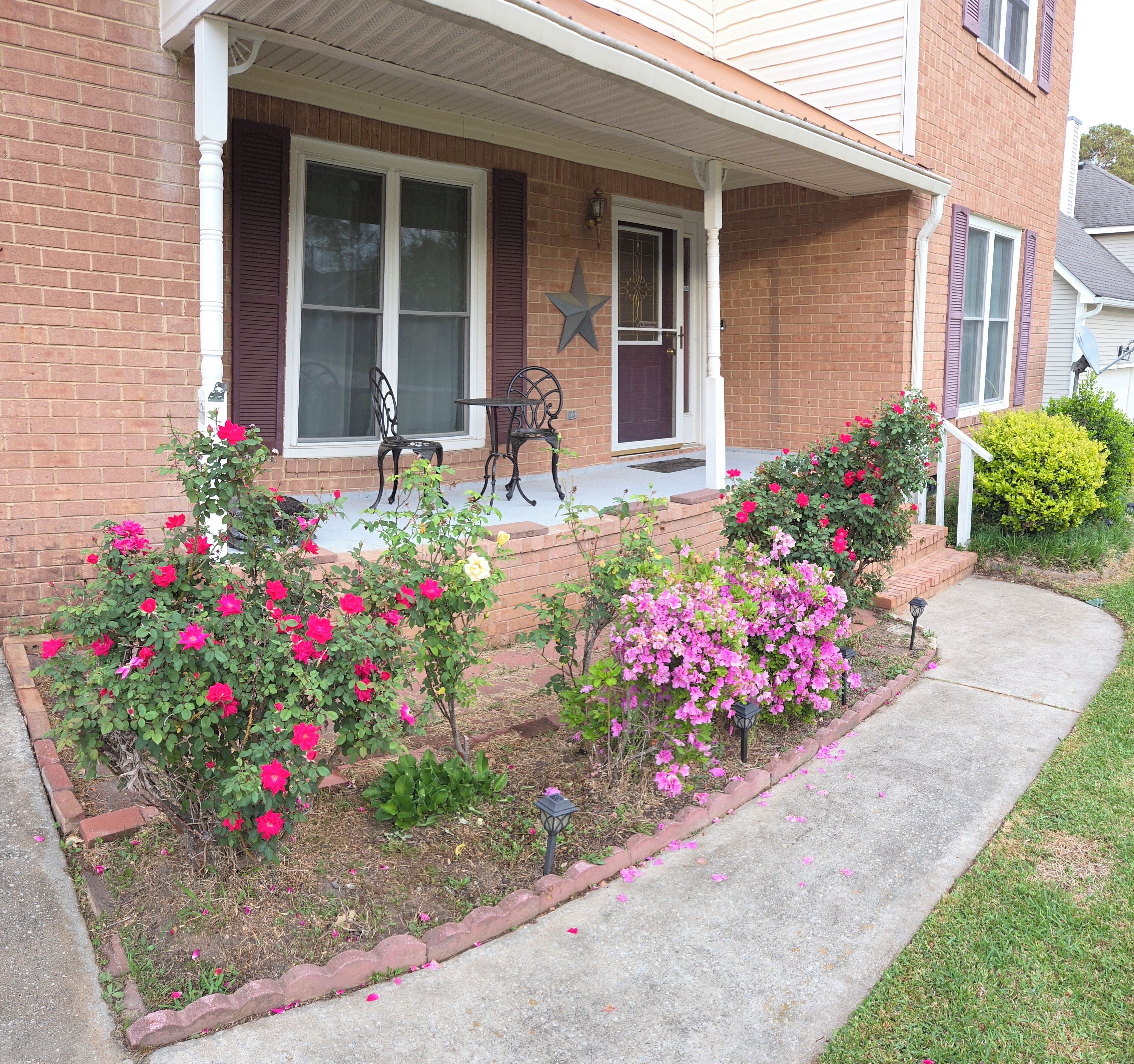 Une maison en briques avec un porche, deux fenêtres, une décoration en forme d’étoile et un jardin avec des fleurs roses.