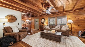 A cozy log cabin interior with a wooden ceiling, a round wall clock, a ceiling fan, a fireplace, and a staircase leading to an upper level. - Maggie Valley Cabin w/ Hot Tub, Fire Pit, Game Room, Sleeps 8, Trailer Friendly (Maggie Valley)