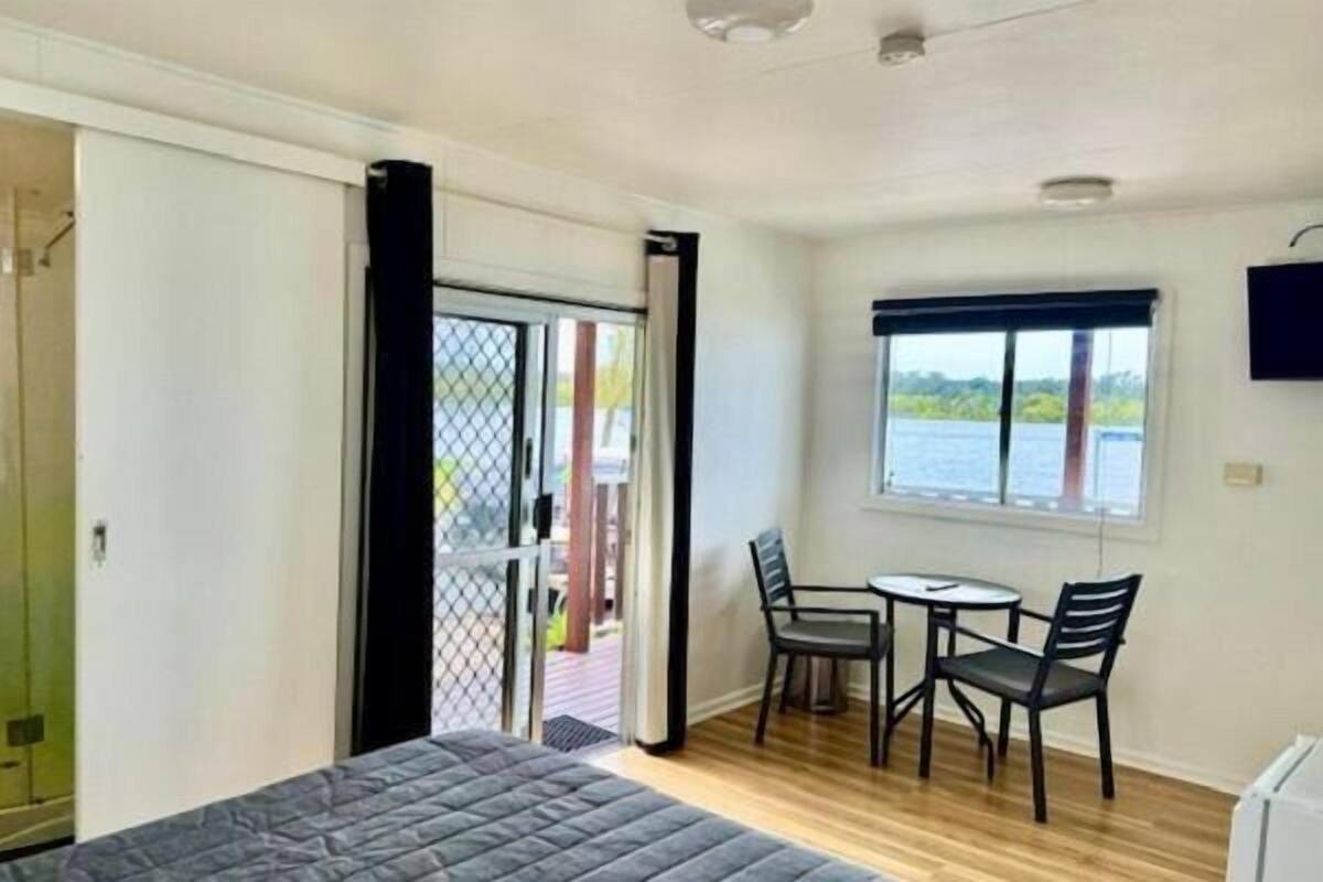 A bedroom with a bed, a shower, a balcony with a table and chairs, and a window with a view of the water.