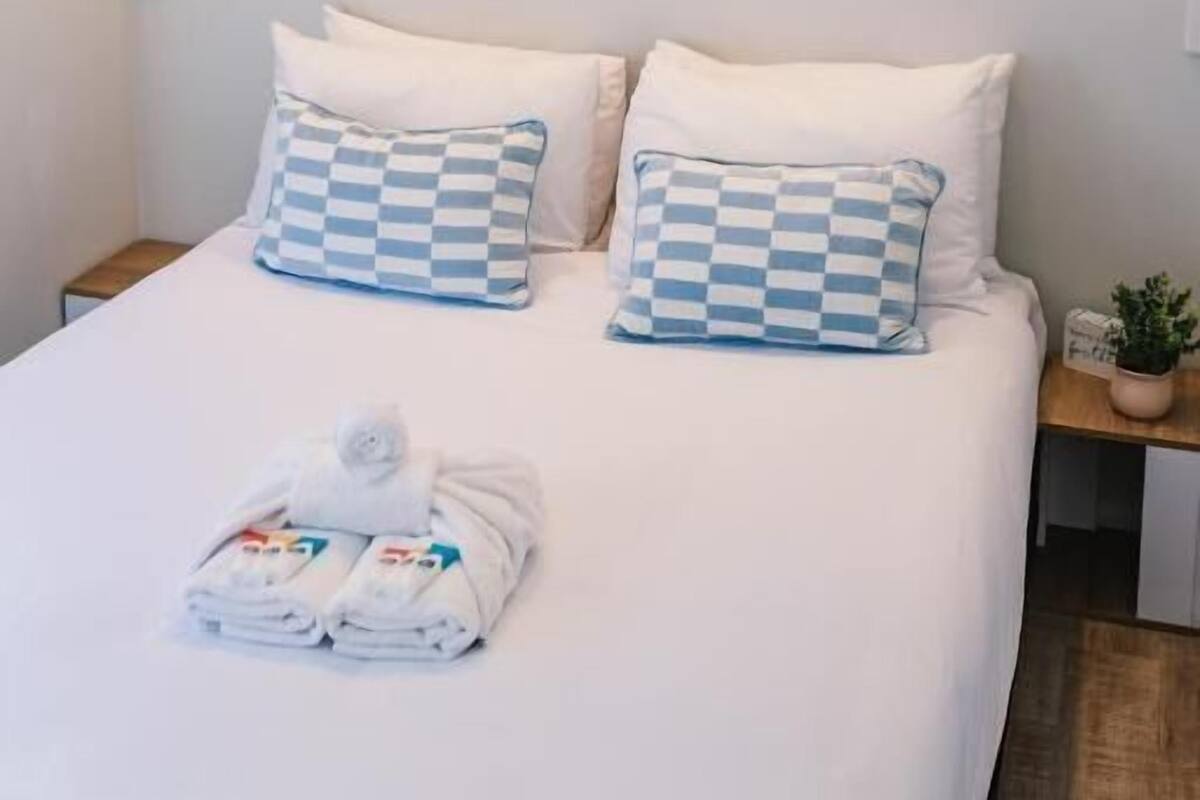 A hotel room with a bed, checkered pillows, a painting of a beach, and a wall-mounted air conditioner.