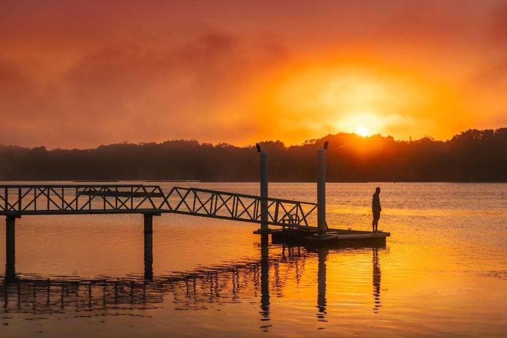 Un muelle se adentra en aguas tranquilas, con un atardecer en el fondo.