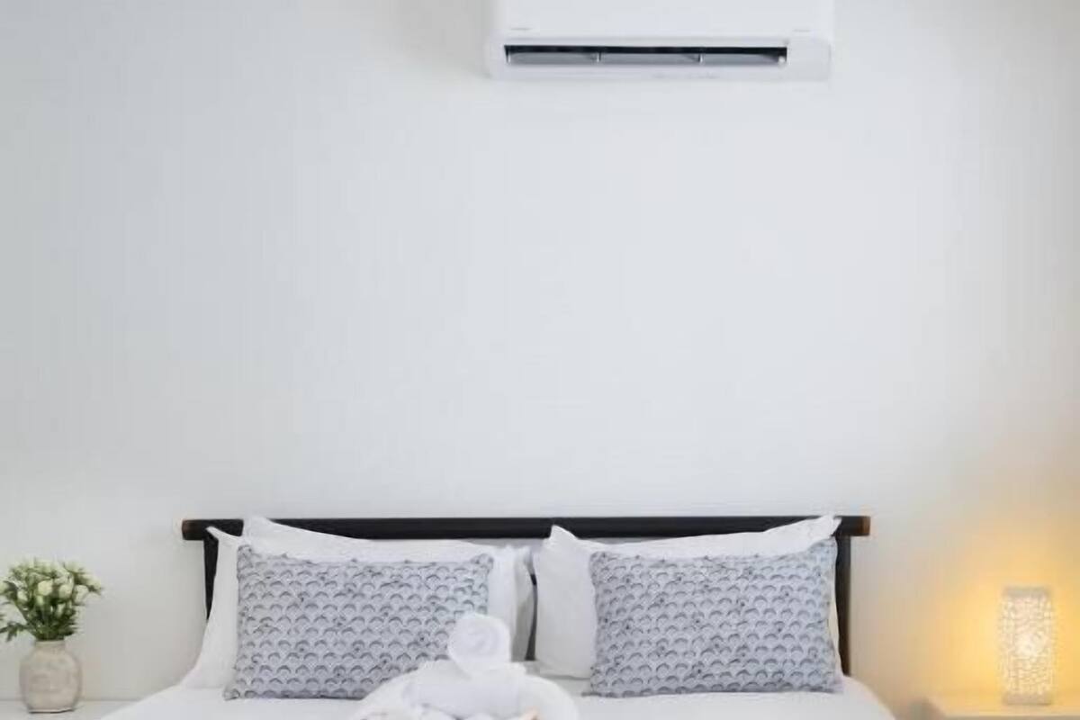 A neatly made bed with white linens, a bedside lamp, and a wall-mounted air conditioner.