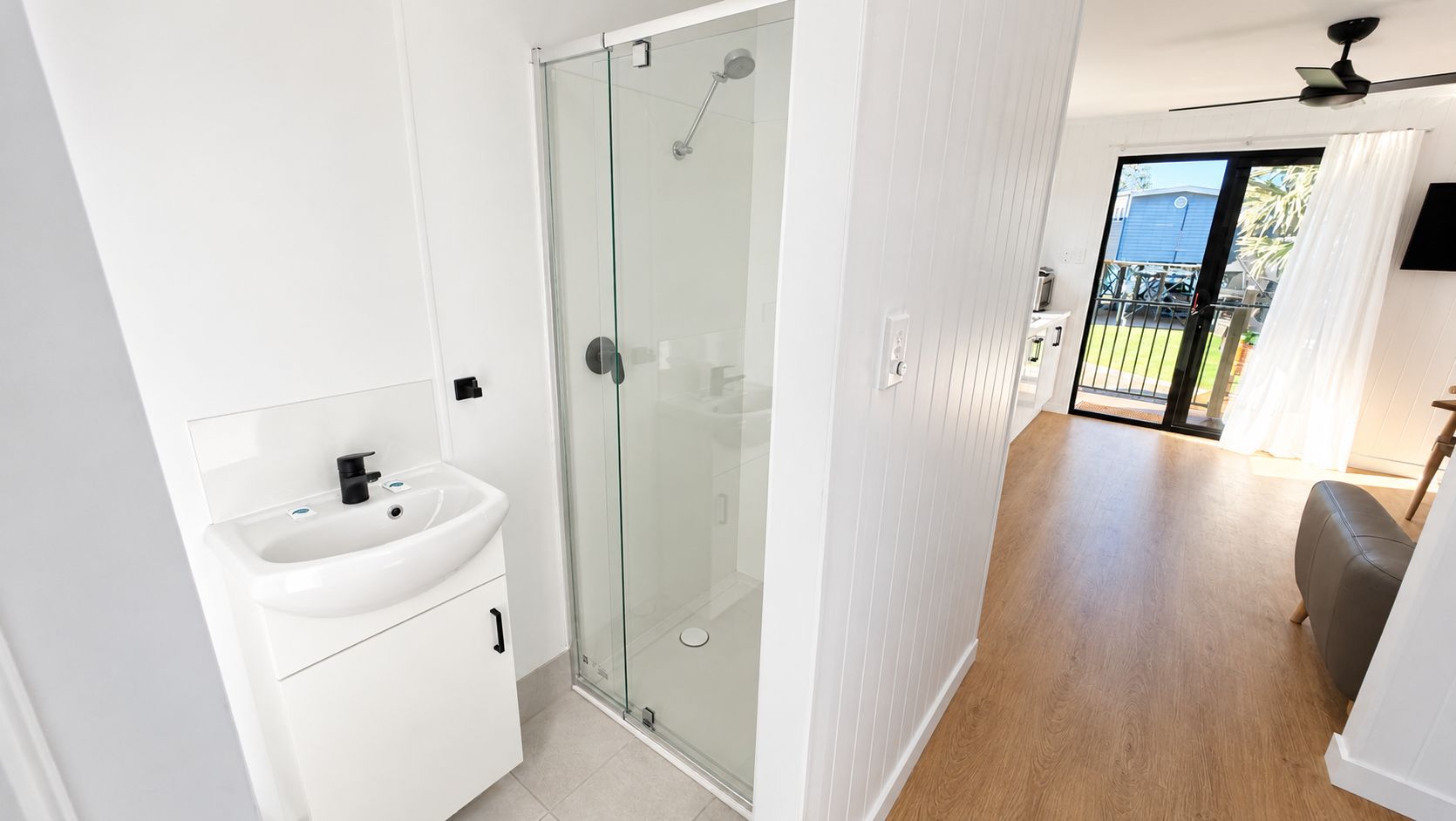 Une salle de bain moderne avec une douche à parois de verre, un lavabo blanc et un plancher en bois donnant sur un balcon.