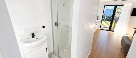Une salle de bain moderne avec une douche à parois de verre, un lavabo blanc et un plancher en bois donnant sur un balcon.