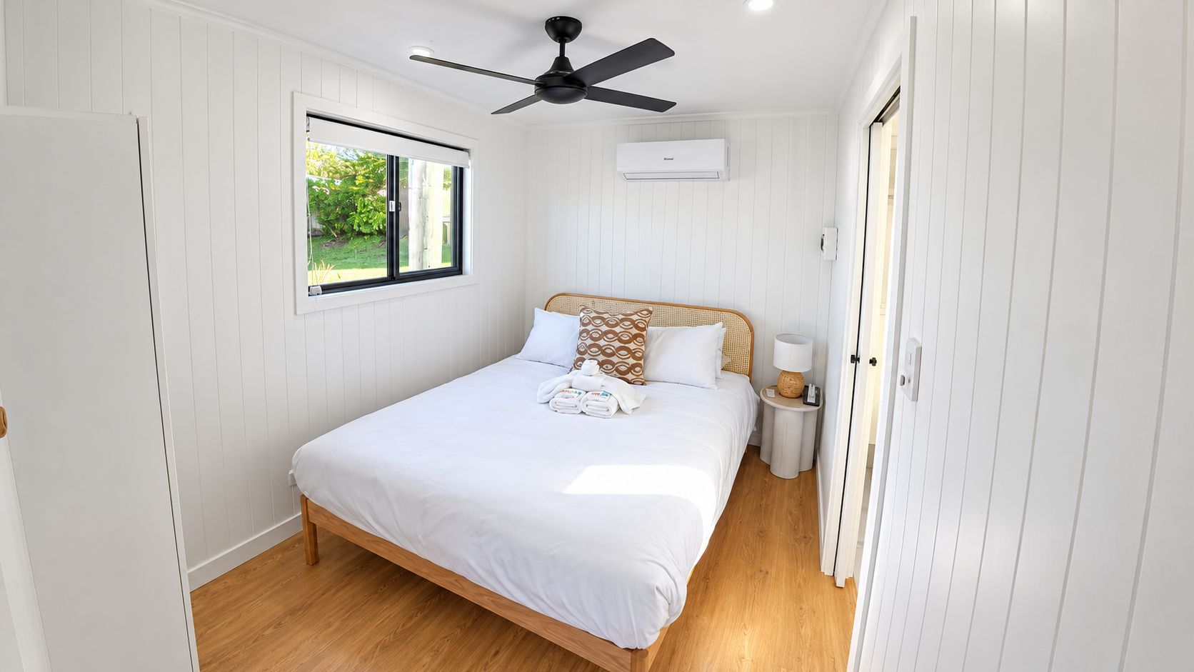 Quarto com cama, ventilador de teto, janela, ar condicionado e mesa de cabeceira com luminária.
