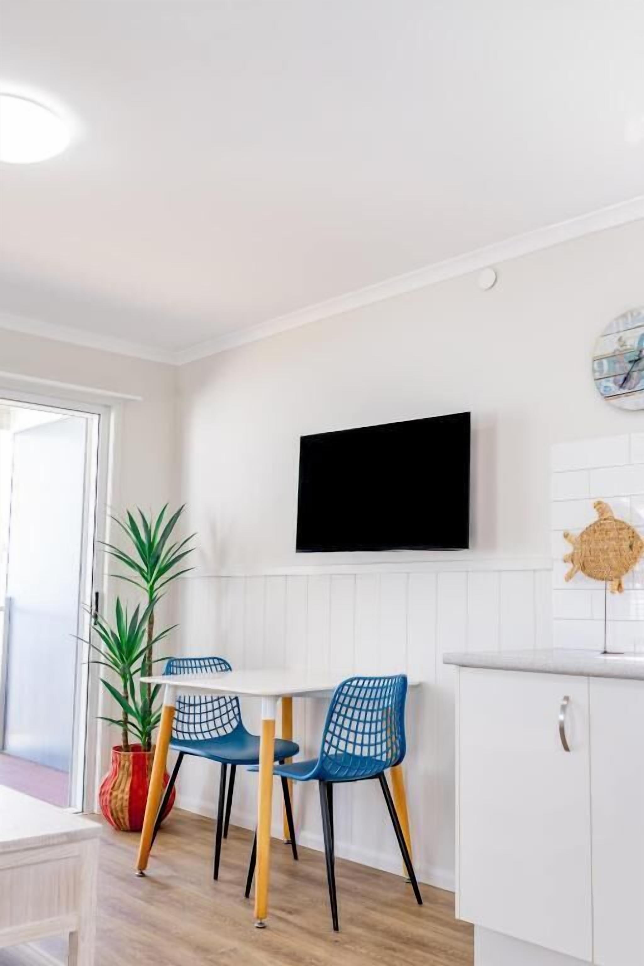 A modern kitchen with a flat-screen TV, white cabinetry, and a dining area with blue chairs.
