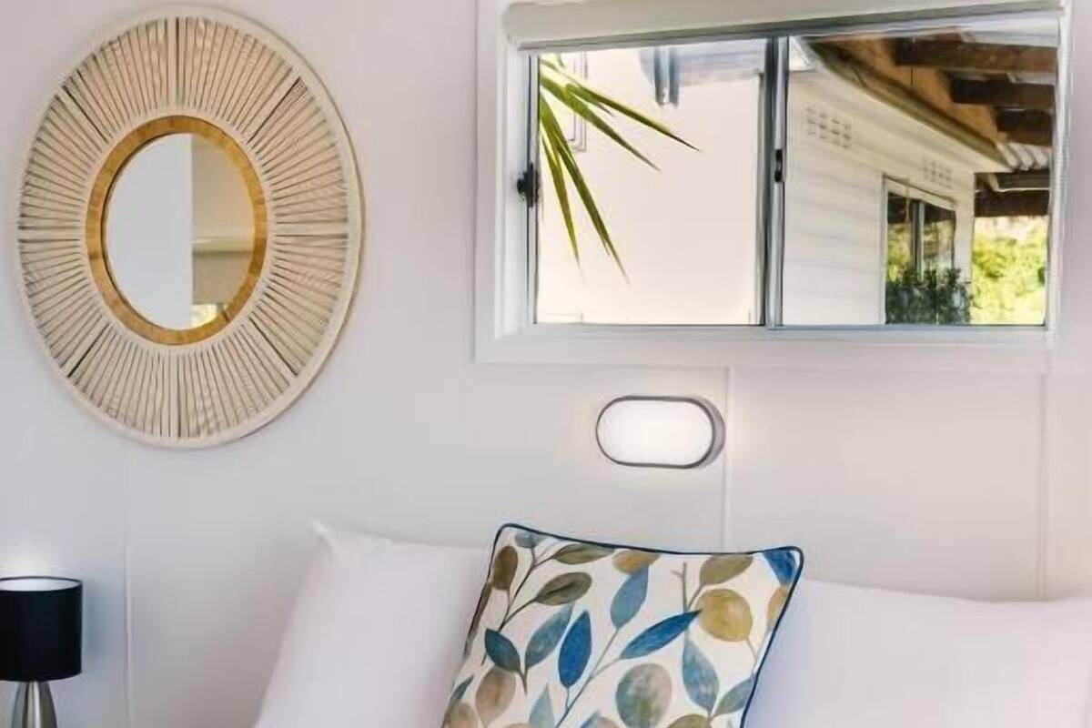 A neatly made bed with a pillow, a round mirror, and a window with a view of greenery.
