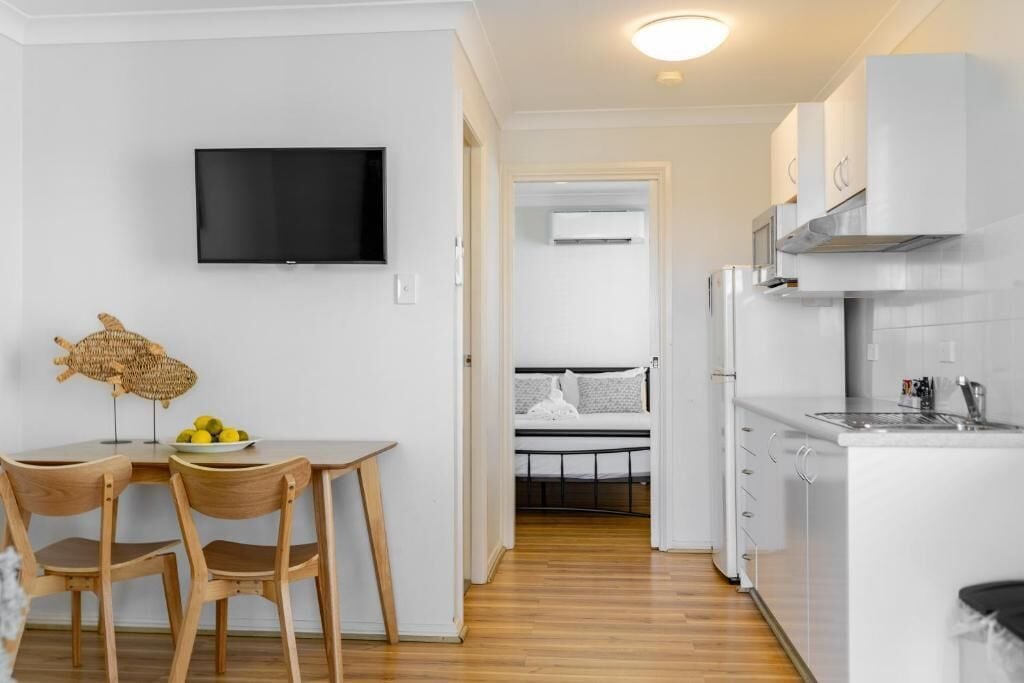 A compact living space with a kitchenette, a dining area with two chairs, a flat-screen TV, and a bed in the background.