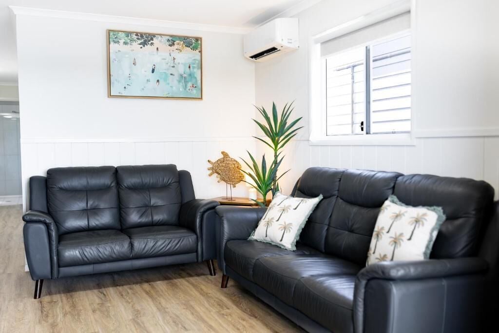 A room with two black leather sofas, a large plant, a painting on the wall, and a window with blinds.