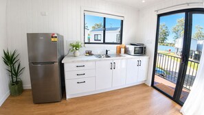 A modern kitchen with a stainless steel refrigerator, white cabinets, a microwave, and a window overlooking a residential area. - Pelican Holiday Park (Nambucca Heads)
