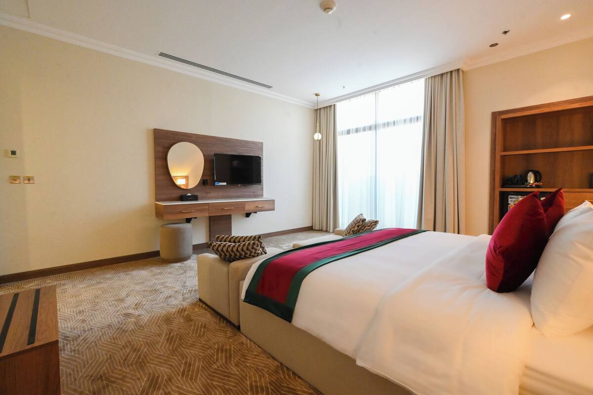 Habitación de hotel con cama, televisor montado en un panel de madera, escritorio, silla y un gran ventanal con cortinas.