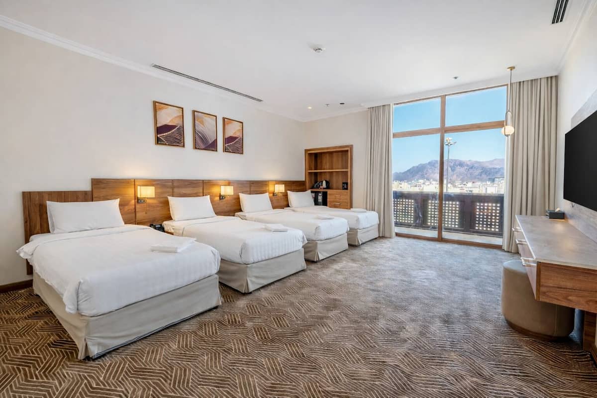 Habitación de hotel con tres camas, un escritorio, un televisor y un ventanal con vistas a la ciudad.