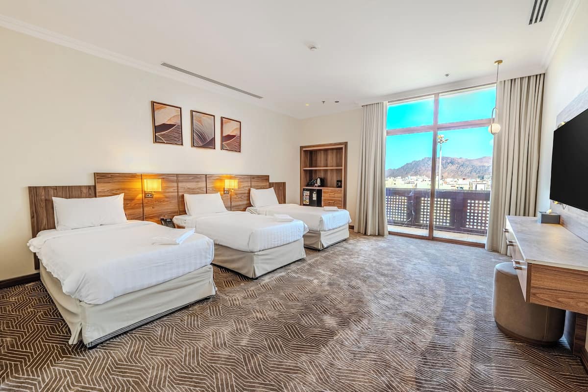 Habitación de hotel con dos camas, un escritorio, una estantería y un ventanal con vista a la ciudad.