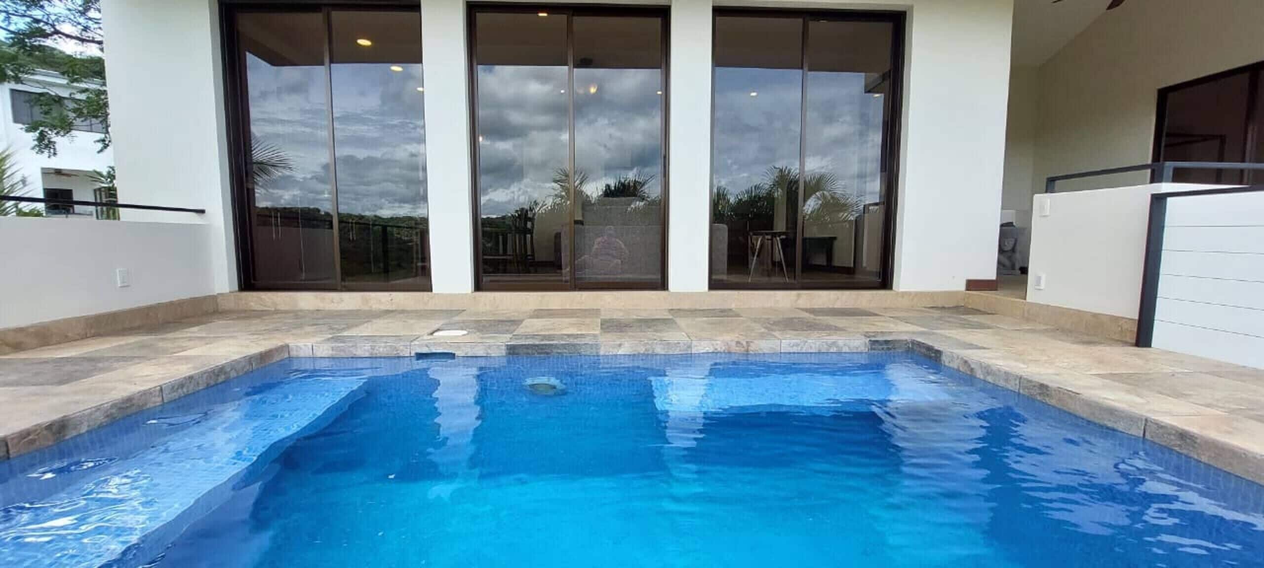 A swimming pool with a tiled deck, large sliding glass doors, and a view of the surrounding area.