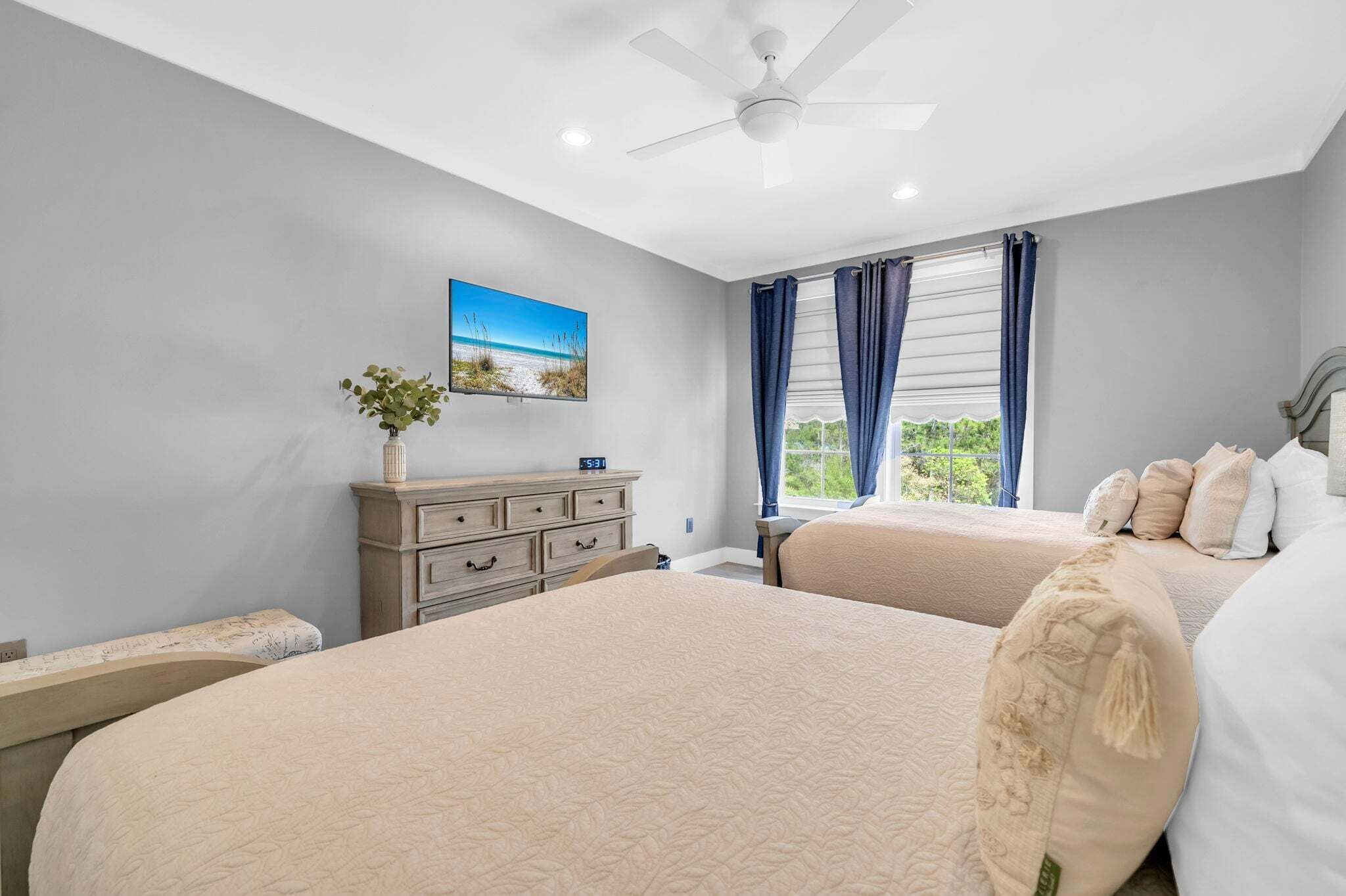 A bedroom with two beds, a dresser, a ceiling fan, a window with curtains, and a painting on the wall.