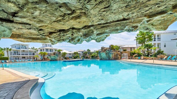 A swimming pool with a rocky overhang, poolside lounge chairs, and a view of residential buildings. - 🦩Book Me Baby! Sea Breeze Escape in Santa Rosa Beach🦩 (Santa Rosa Beach)