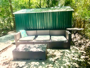 A covered outdoor seating area with a sofa and ottoman, surrounded by greenery. - Cozy Home: Fire Pit+Pets: near Riverside+Downtown (Jacksonville)