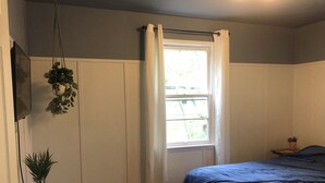 A bedroom with a bed, a window with curtains, a potted plant on a nightstand, and a hanging plant. - Cozy Home: Fire Pit+Pets: near Riverside+Downtown (Jacksonville)