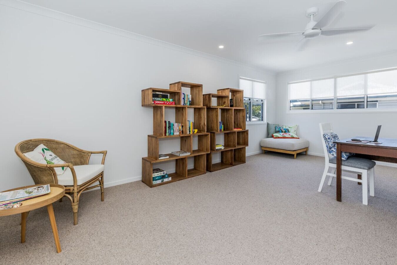A room with a large wooden bookshelf, a wicker chair, a small round table, a sofa, and a desk with a laptop.
