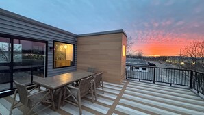 Una terraza con mesa y sillas, un edificio moderno con amplios ventanales y vistas al atardecer.