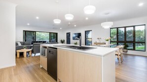 A modern kitchen with a central island, a dining area with a table and chairs, and large windows offering a view of the outdoors. - Coastal Haven - Agnes Water (Agnes Water)