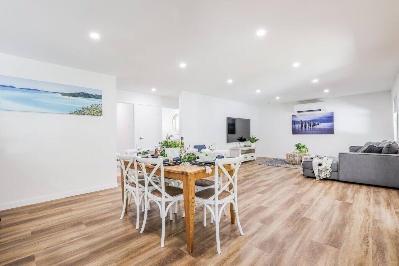 A modern living room with a dining area, wooden flooring, white walls, and a flat-screen TV.