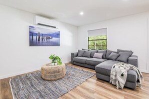 A modern living room with a grey sectional sofa, a round wicker ottoman, a large abstract painting, and a window with a view of greenery. - Ocean Haze - Prime Beachfront Getaway! (Agnes Water)