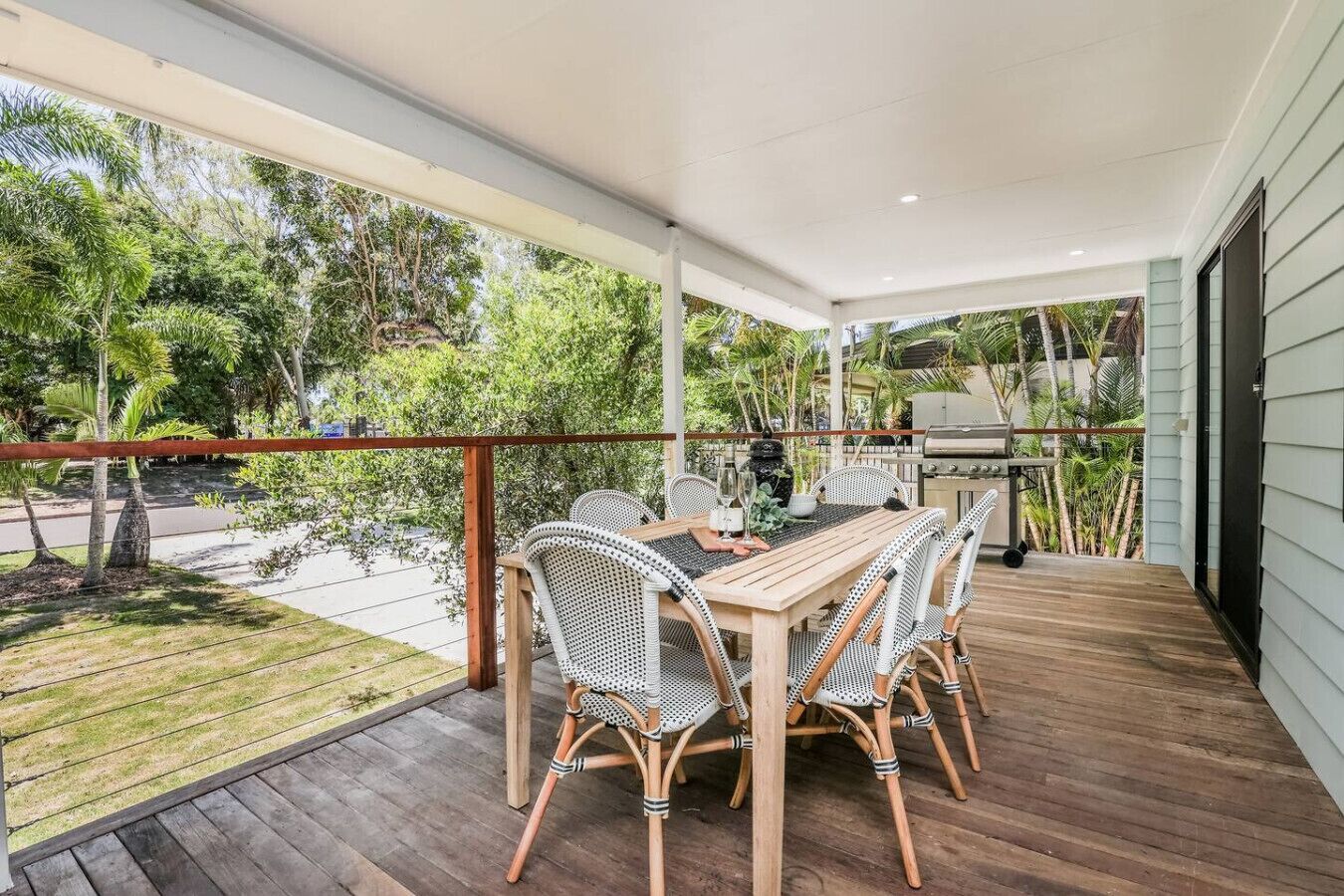 An outdoor patio with a wooden table and wicker chairs, a grill, and a view of greenery.