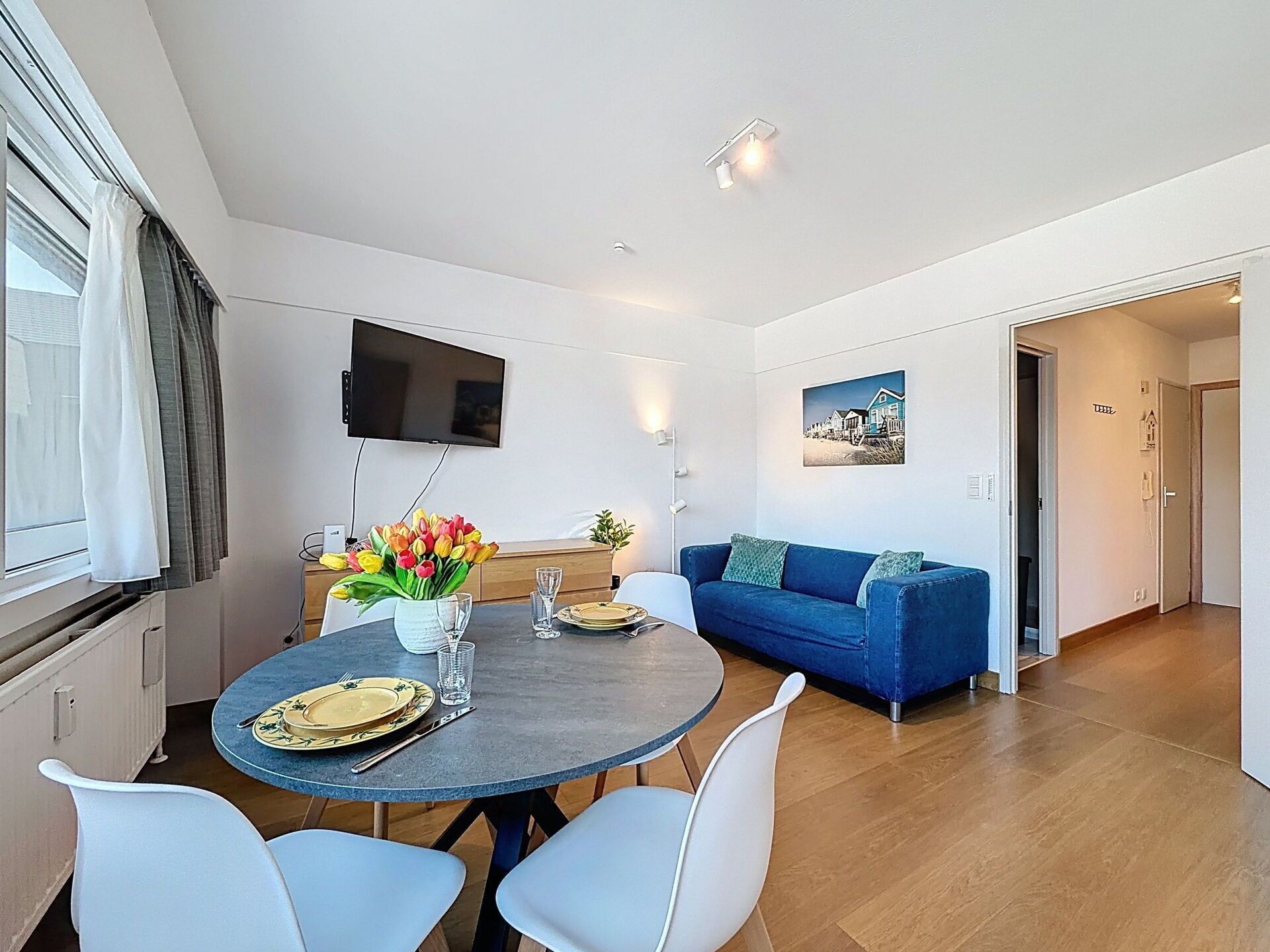 Een moderne woonkamer met een blauwe bank, een eettafel met stoelen, een flatscreen televisie aan de wand en een schilderij.