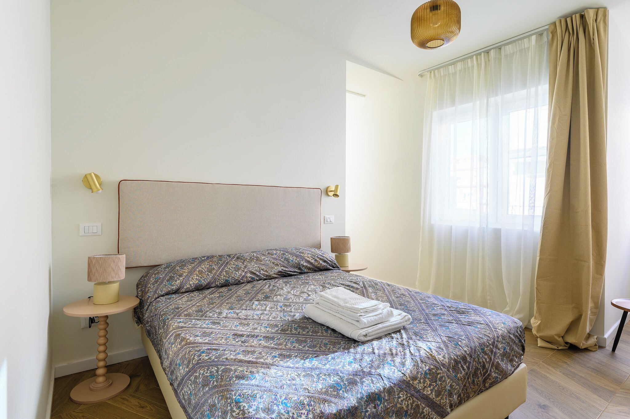 A neatly made bed with patterned bedding, a bedside lamp, and a window with sheer curtains.