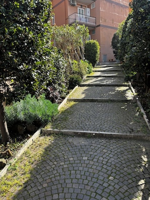 A cobblestone pathway flanked by lush greenery and a building with a balcony. - Spacious bright and cozy penthouse with terrace close to metro station (Roma)